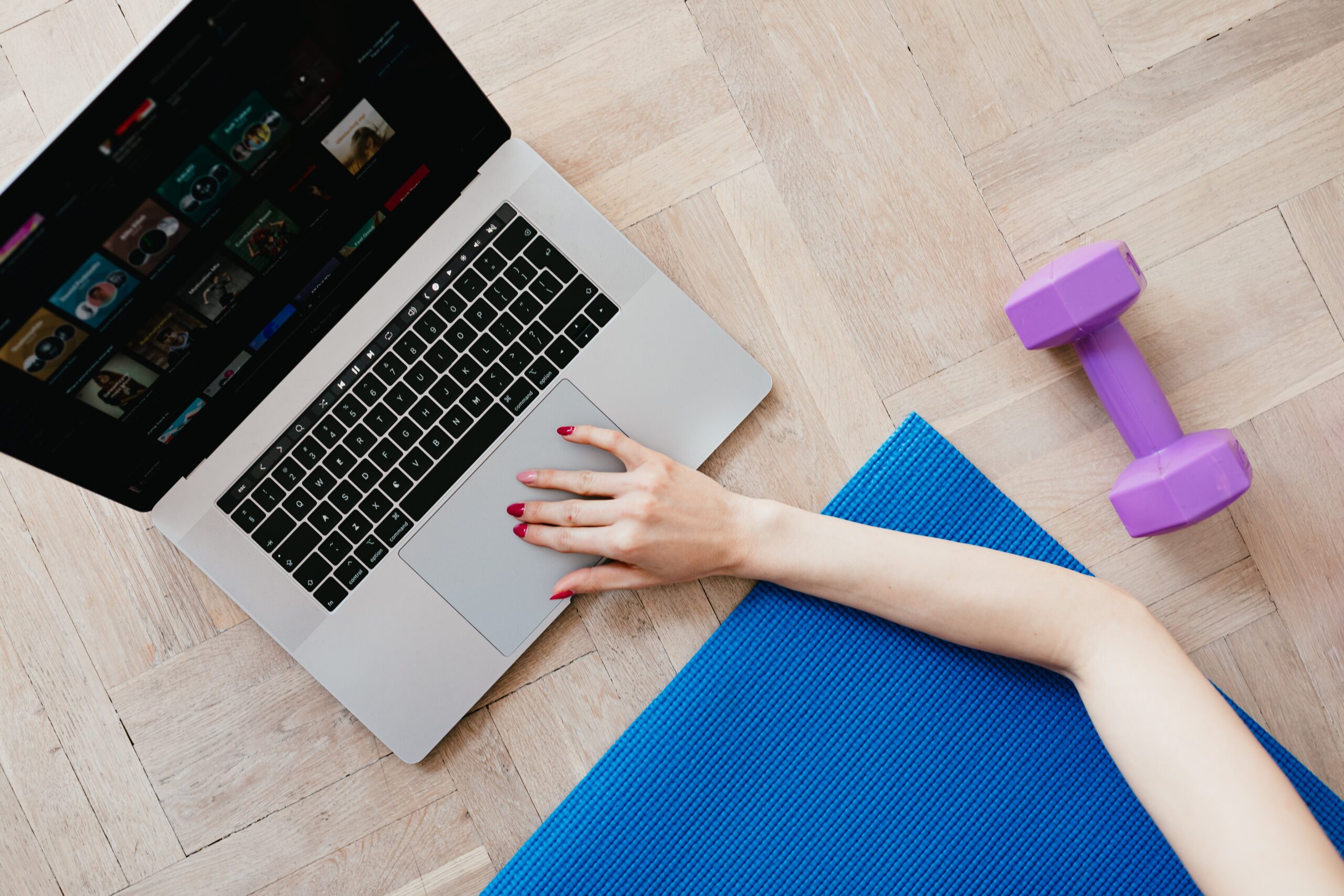 Top view of crop anonymous female looking for video workout courses on laptop while sitting on blue yoga mat with purple dumbbell beside on parquet floor
